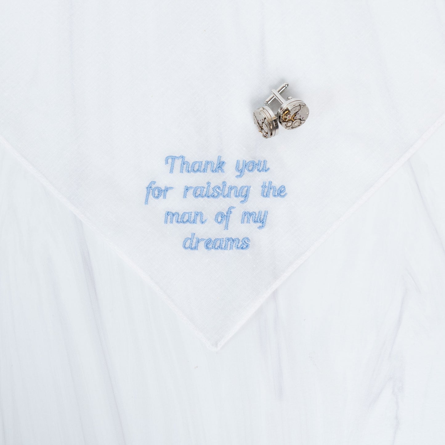 A white handkerchief with light blue embroidered text reading 'Thank you for raising the man of my dreams' and a pair of silver cufflinks placed on top.