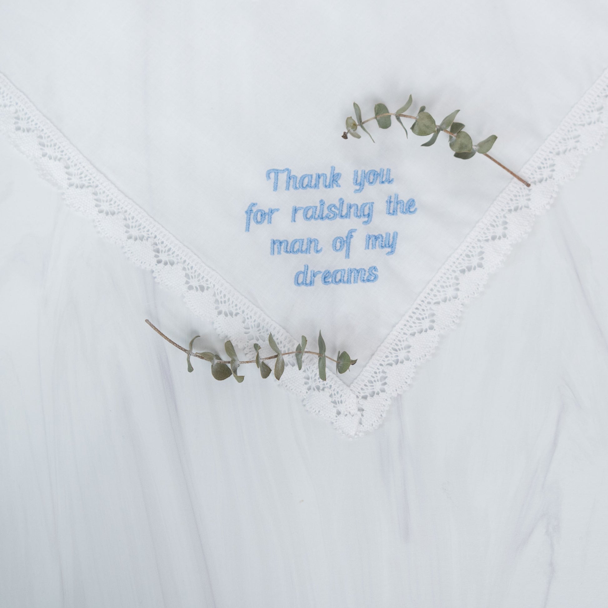 A white lace-trimmed handkerchief with light blue embroidered text reading 'Thank you for raising the man of my dreams,' styled with two small eucalyptus sprigs.