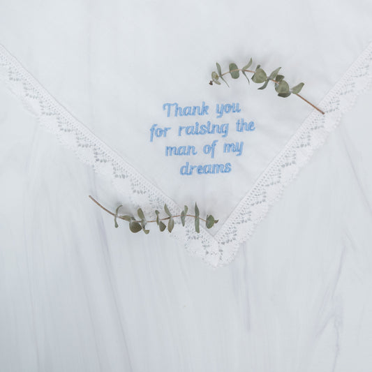 A white lace-trimmed handkerchief with light blue embroidered text reading 'Thank you for raising the man of my dreams,' styled with two small eucalyptus sprigs.