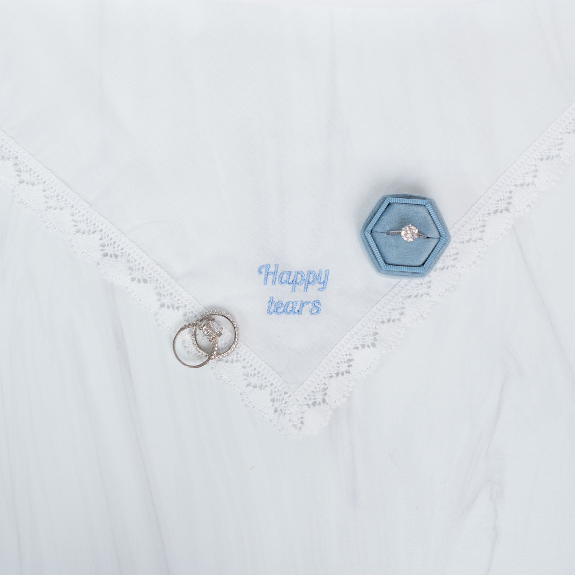 A close-up of a white lace-trimmed handkerchief laid flat, with a small blue hexagonal ring box holding a ring and a pair of silver rings placed on the fabric. Blue embroidered text reads 'Happy tears'.