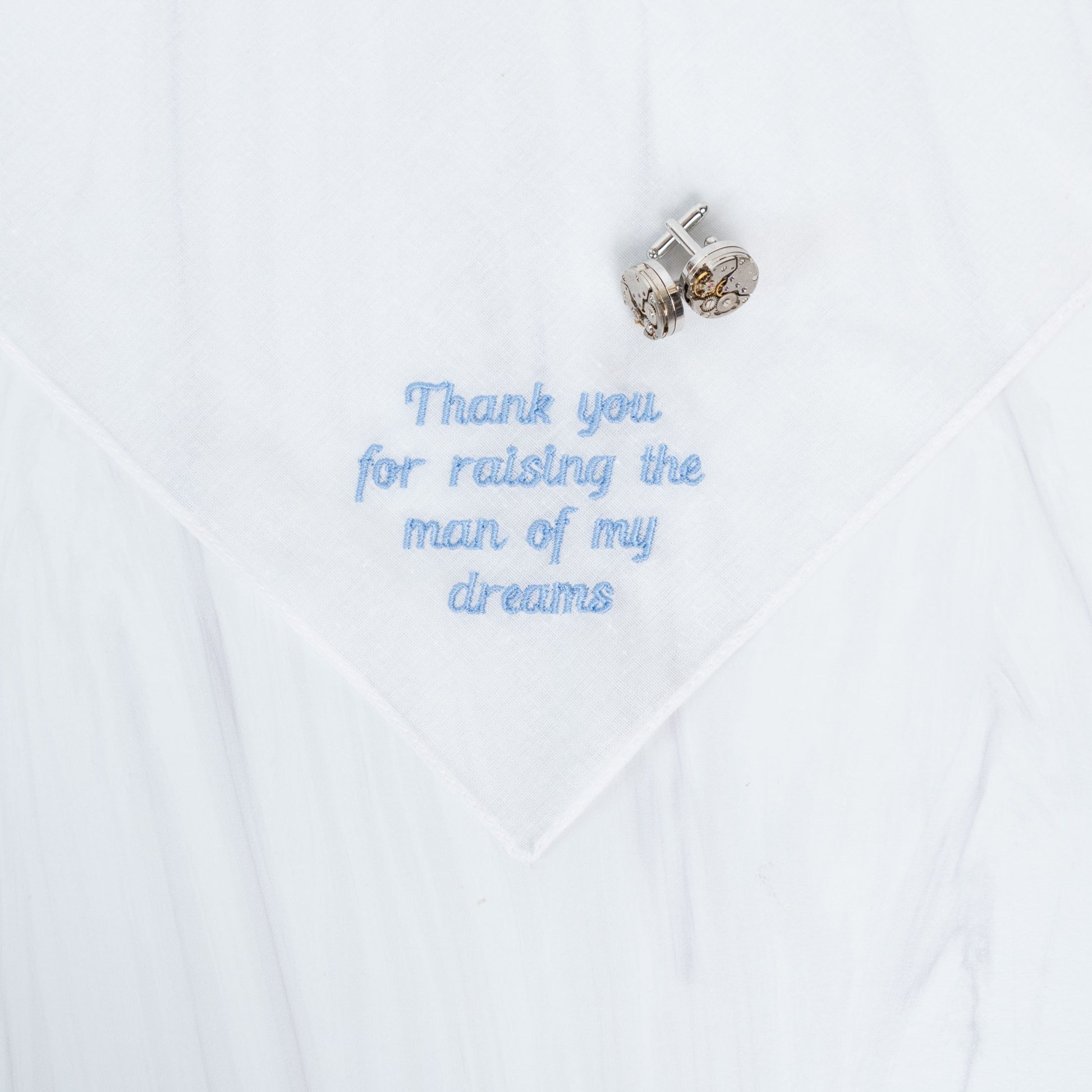 A white handkerchief with light blue embroidered text reading 'Thank you for raising the man of my dreams' and a pair of silver cufflinks placed on top.