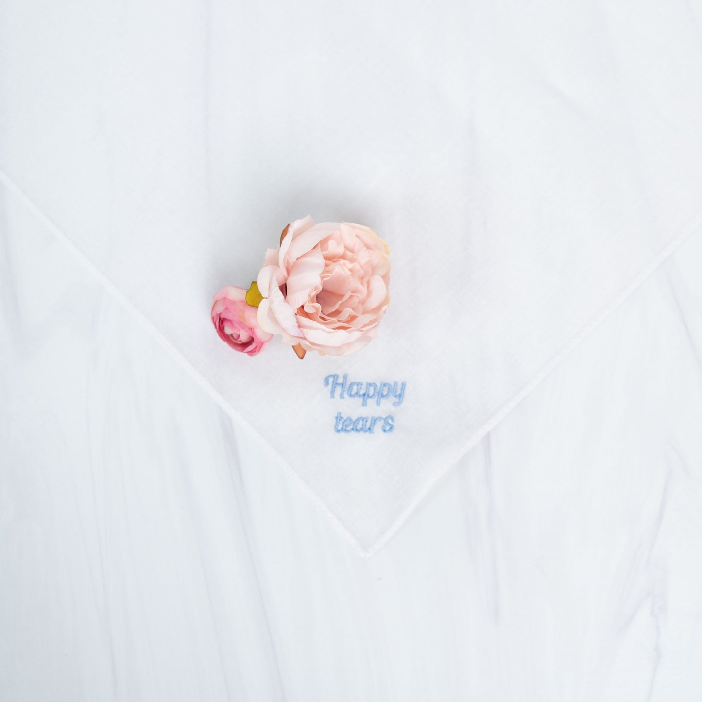 A white square handkerchief with embroidered blue text reading 'Happy tears' and a small pale pink flower placed on it, photographed on a white fabric background.