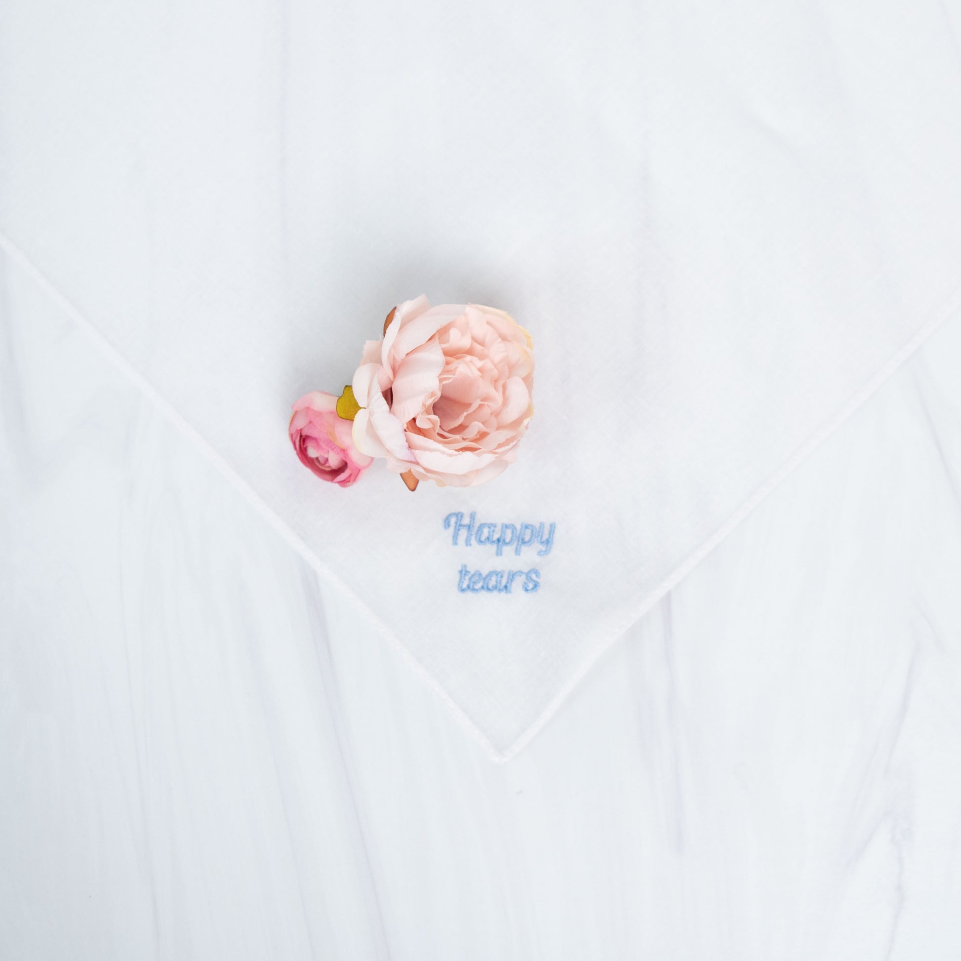 A white square handkerchief with embroidered blue text reading 'Happy tears' and a small pale pink flower placed on it, photographed on a white fabric background.