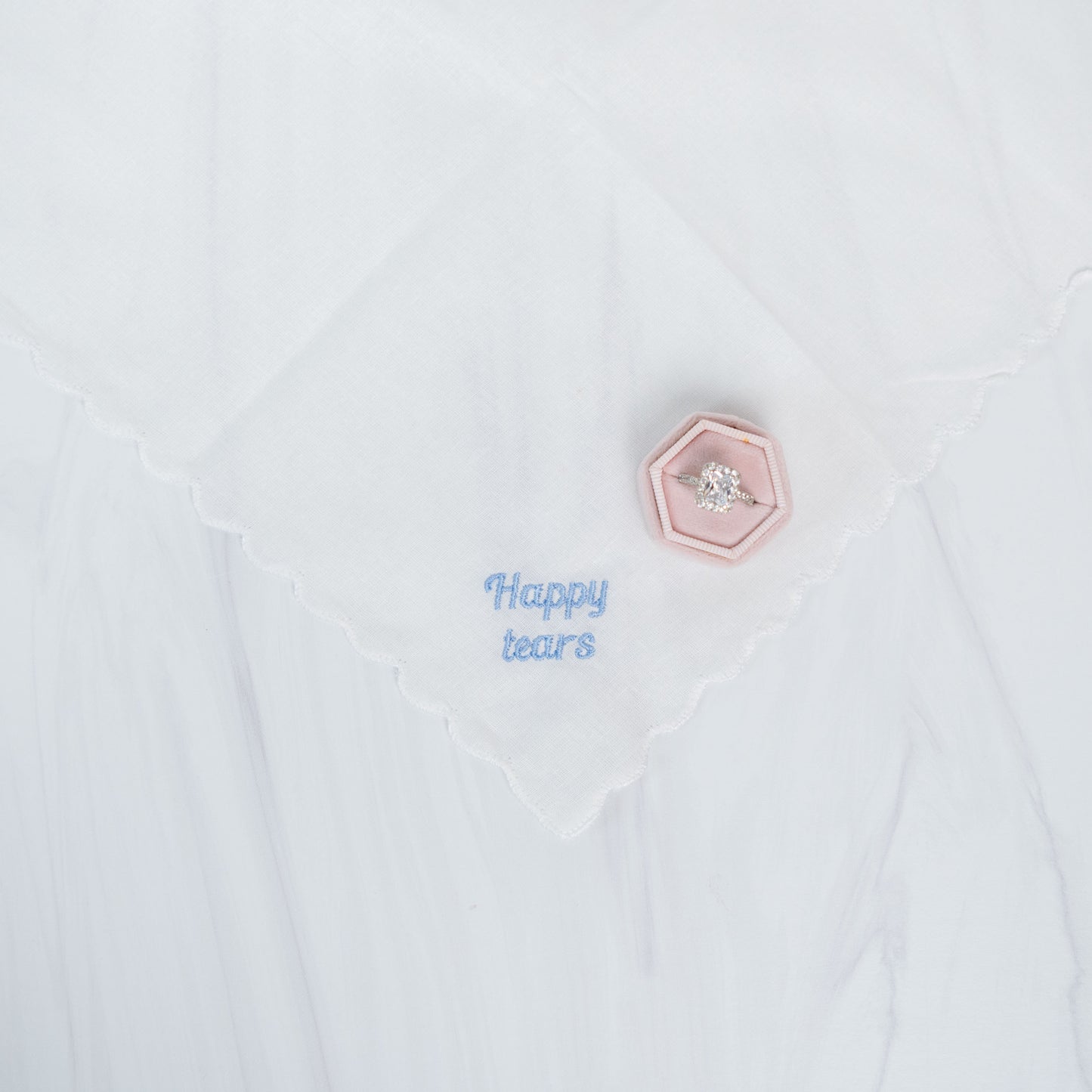 A close-up of a white handkerchief with a scalloped edge, featuring light blue embroidered text reading 'Happy tears' and a small pink hexagonal pin with a clear rhinestone.
