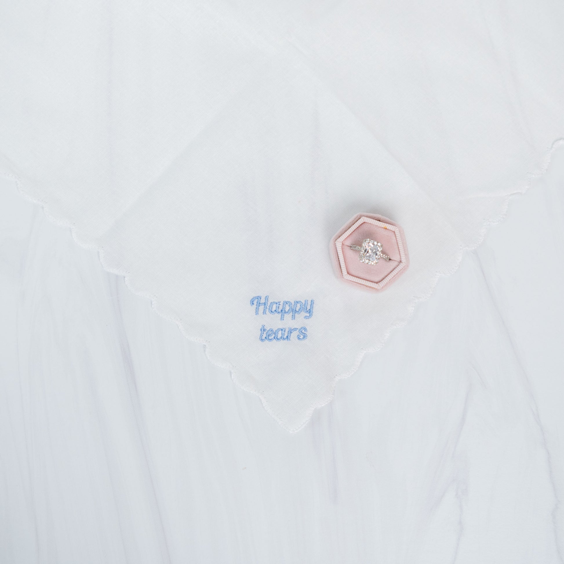 A close-up of a white handkerchief with a scalloped edge, featuring light blue embroidered text reading 'Happy tears' and a small pink hexagonal pin with a clear rhinestone.