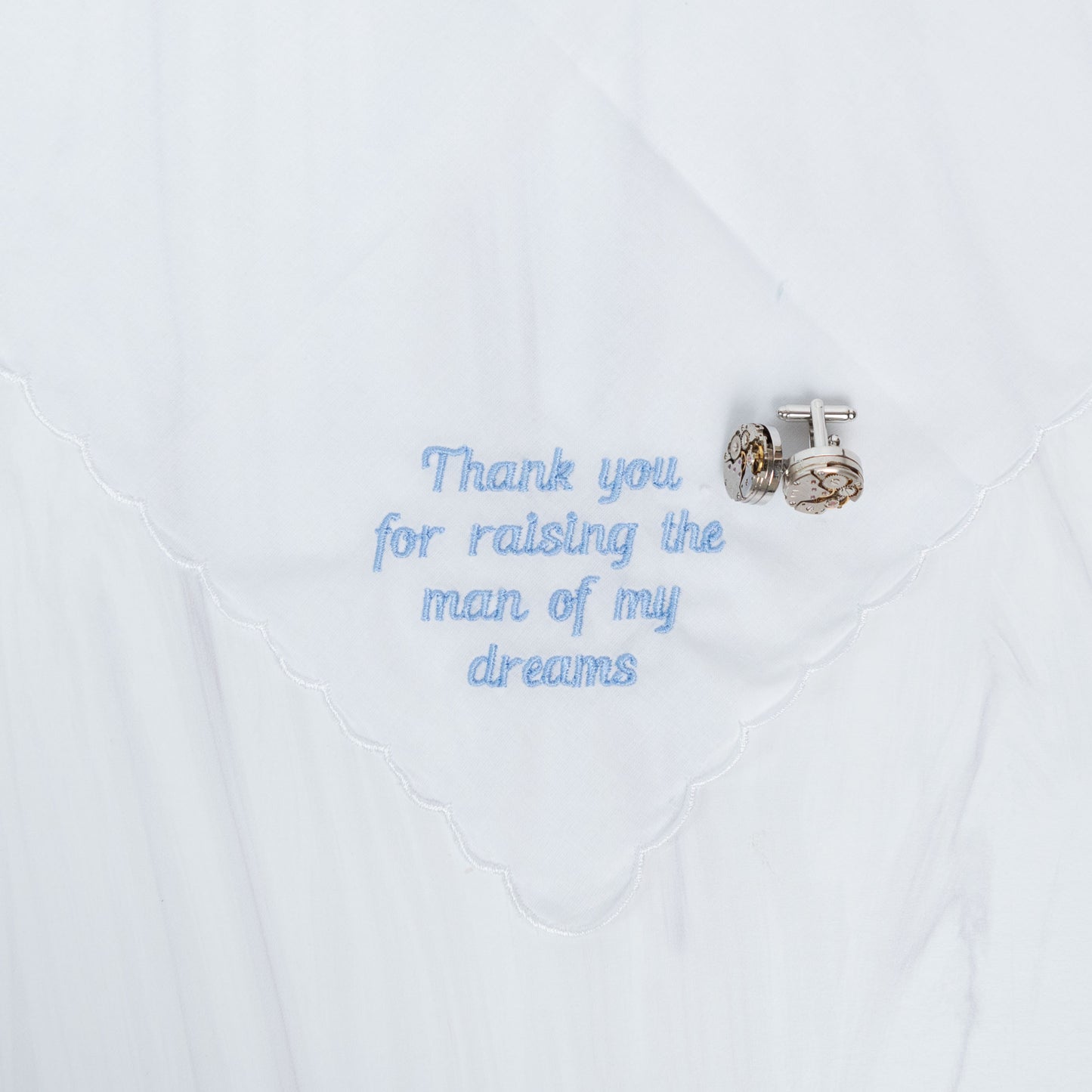 A white embroidered handkerchief with scalloped edges, featuring the phrase 'Thank you for raising the man of my dreams' in light blue script. A silver-toned cufflink is pinned to the right side of the embroidery.