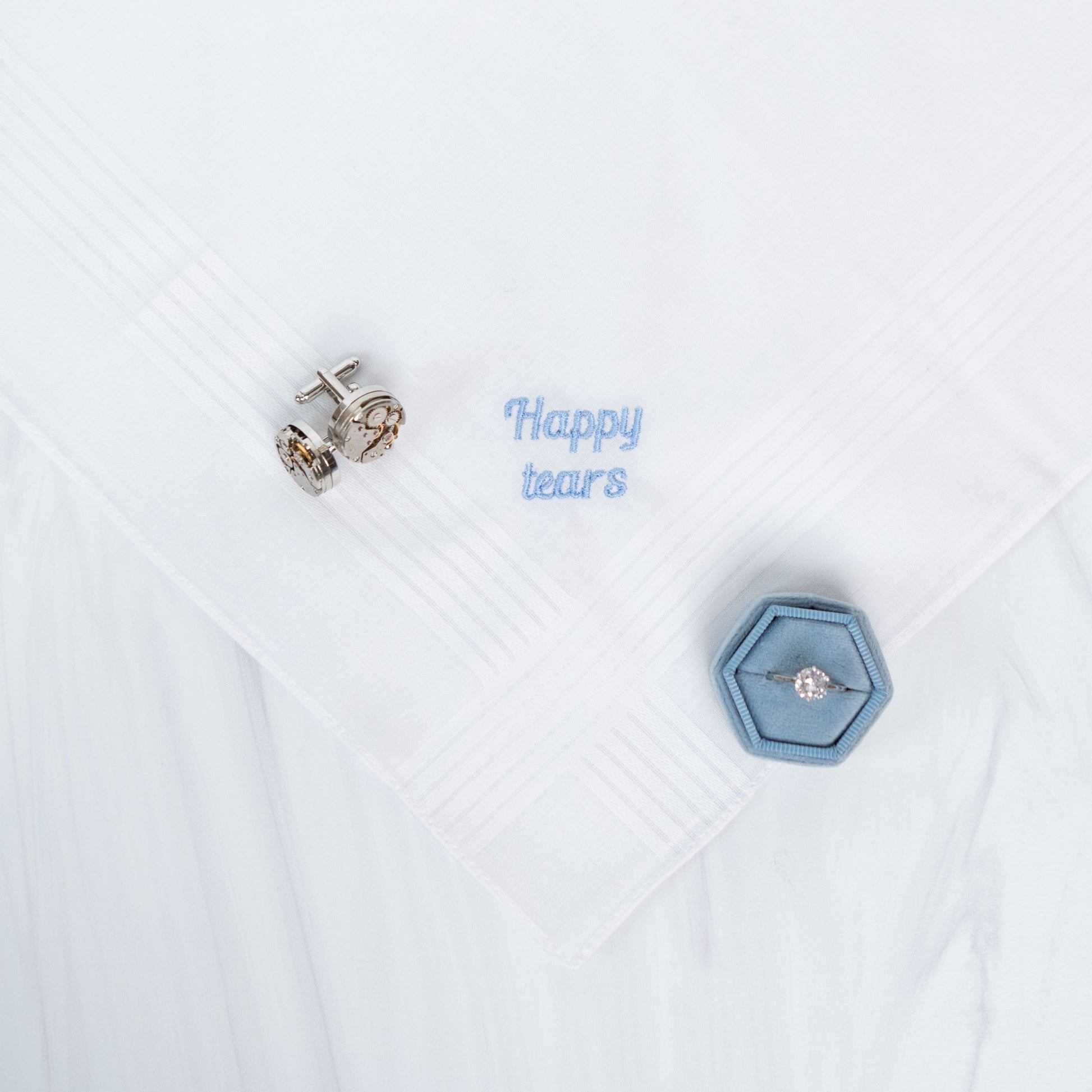 Silver-toned cufflinks displayed on a white handkerchief next to a small blue hexagonal ring box containing a ring and the embroidered text 'Happy tears'.