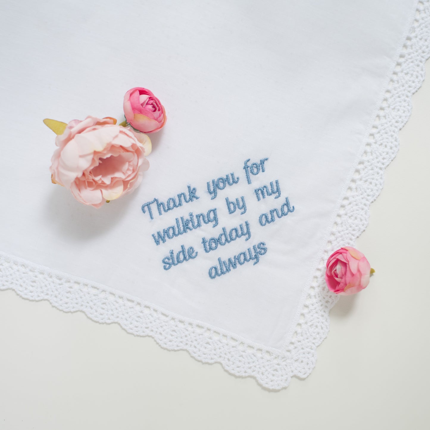 A white lace-trimmed handkerchief with blue embroidered text reading 'Thank you for walking by my side today and always,' styled with pink artificial flowers on a light neutral background.