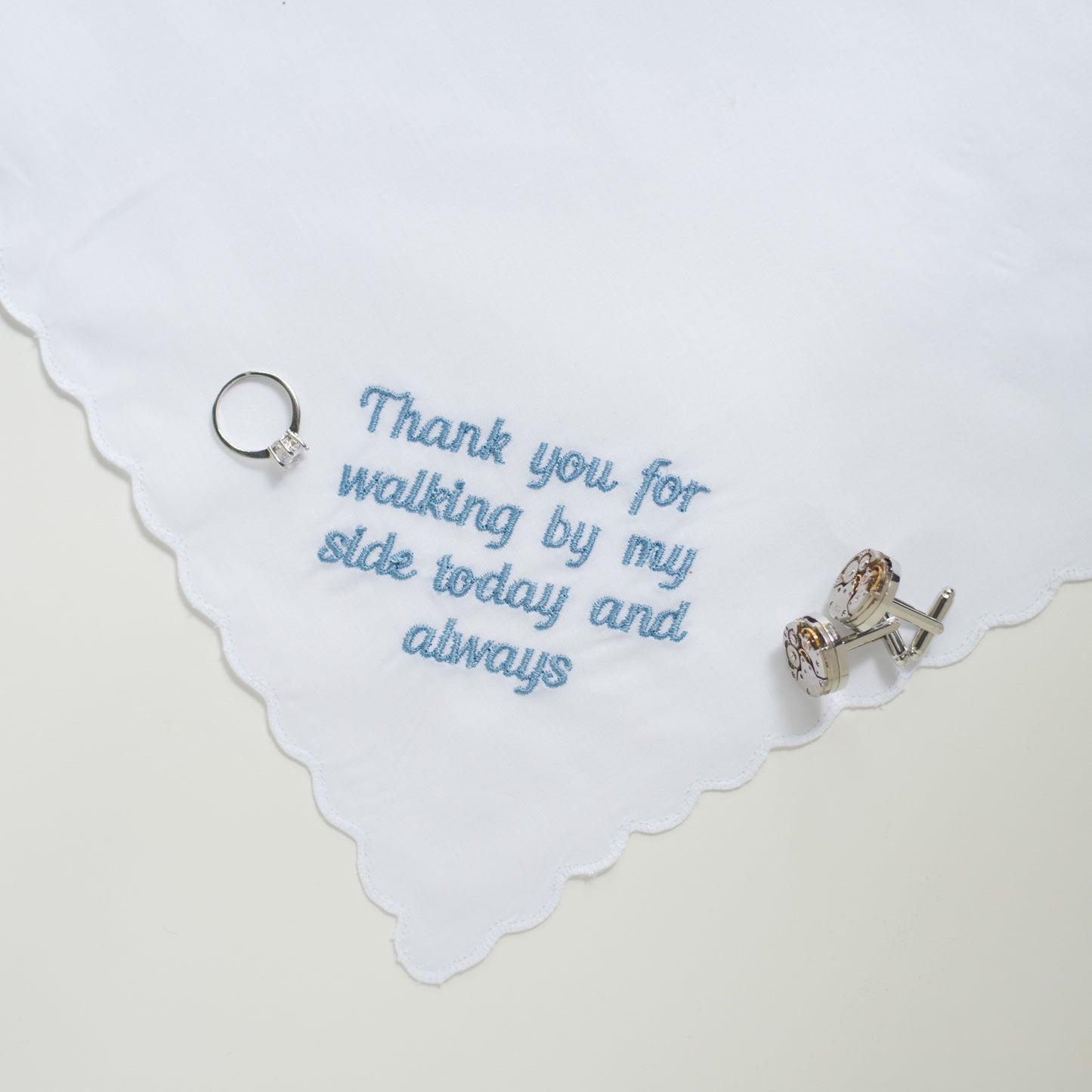 A white handkerchief with scalloped edges, embroidered with the phrase 'Thank you for walking by my side today and always' in light blue script. A silver ring and a silver cufflink are placed on the fabric as styling props against a warm off-white background.