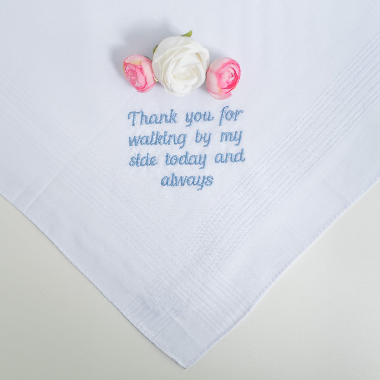 A white square handkerchief with light blue embroidered text reading 'Thank you for walking by my side today and always', styled with three small artificial roses on a light beige surface.